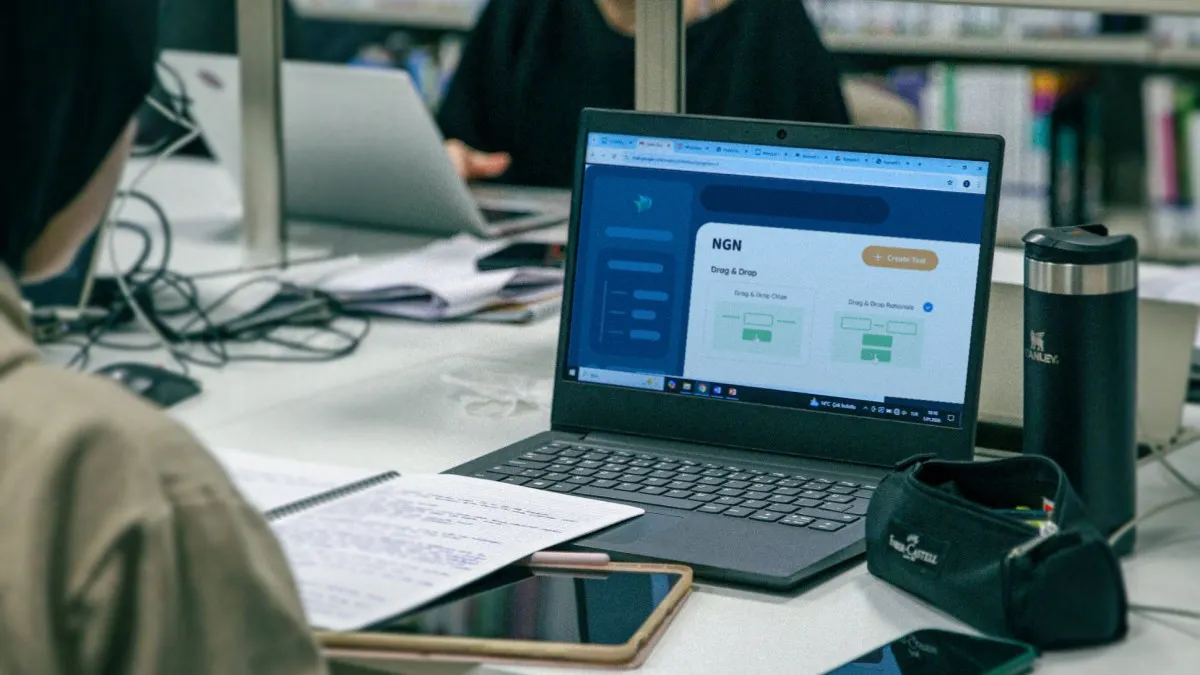 Student studying on a laptop in a library with notes and a tablet while preparing for NGN questions